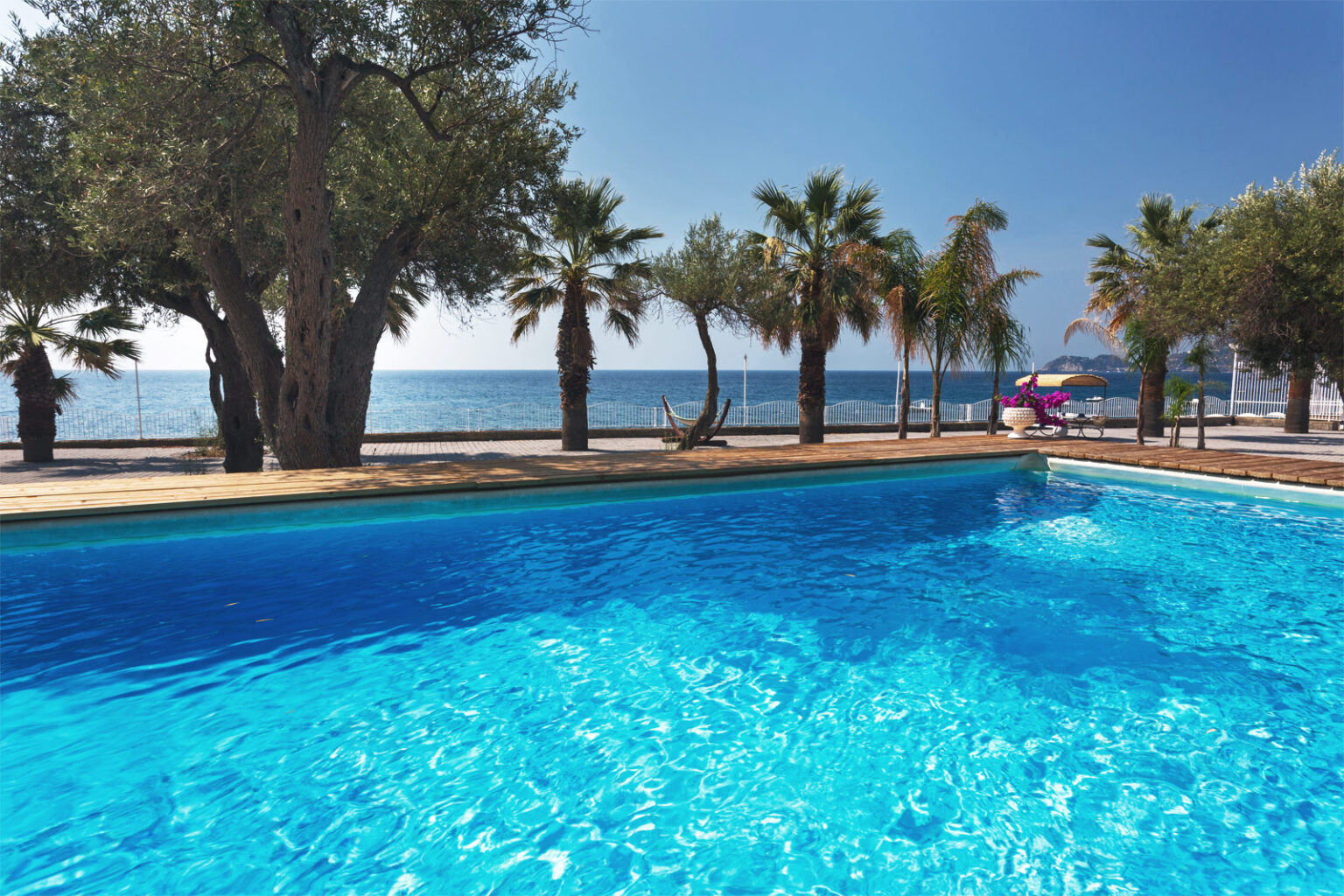 Scenic view of a sparkling blue swimming pool surrounded by palm trees, with a serene ocean and clear blue sky in the background, ideal for a tropical vacation setting. | Only on SopranoVillas