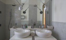 Modern bathroom design featuring a double vanity with two white vessel sinks, stylish wall-mounted faucets, and contemporary lighting. The space includes a walk-in shower with sleek fixtures and a minimalist aesthetic, complemented by mosaic tiles and a bright, airy feel.