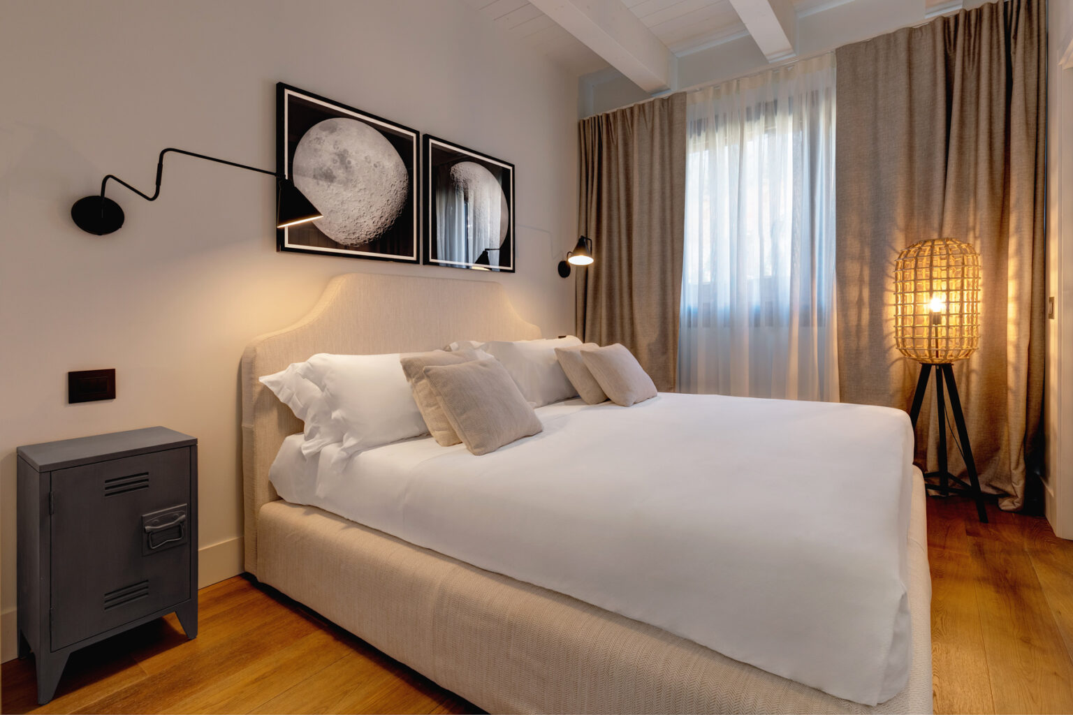 Modern bedroom featuring a comfortable bed with decorative pillows, stylish wall art, a sleek nightstand, and warm lighting from a floor lamp, complemented by soft curtains and wooden flooring. | Only on SopranoVillas