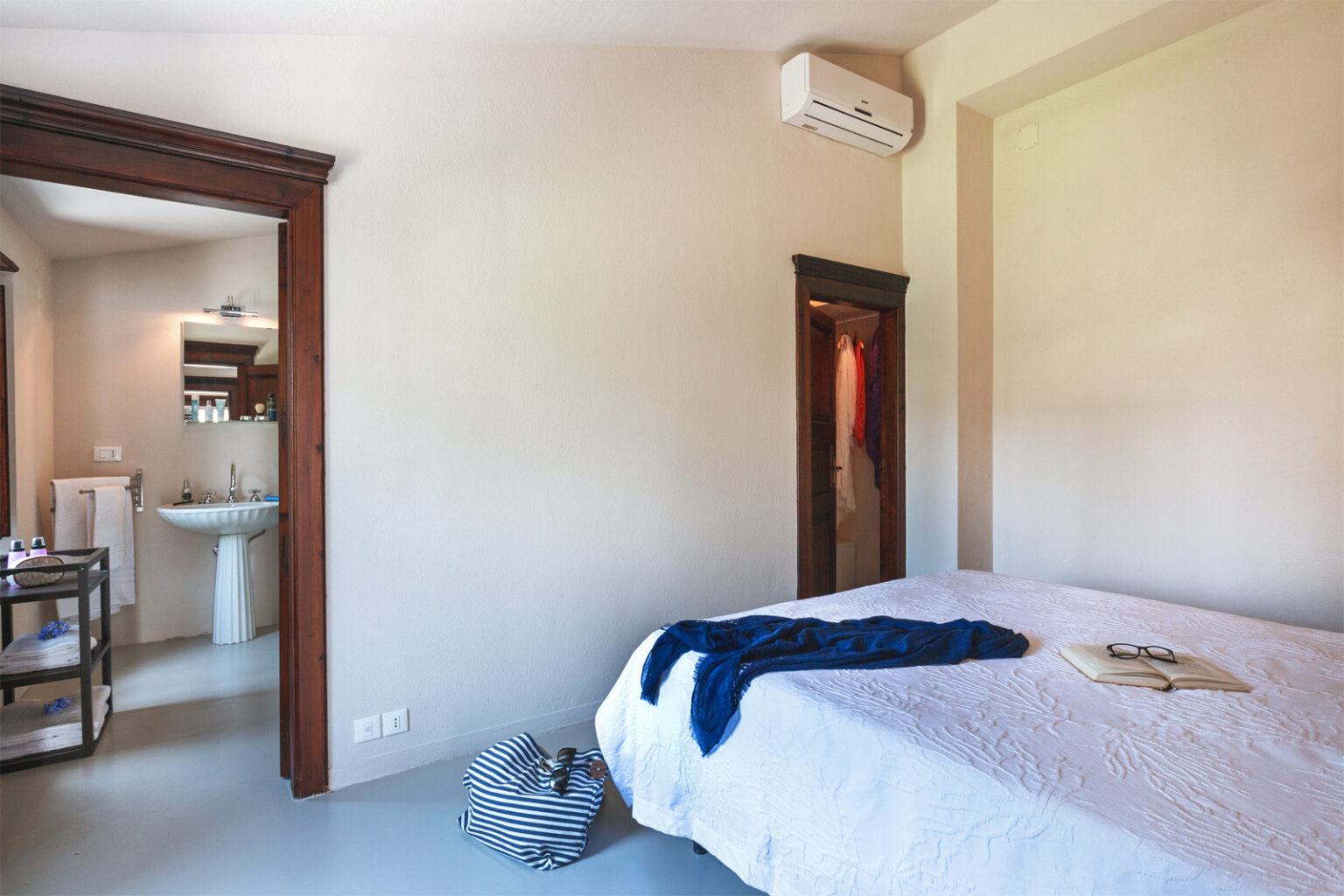 Modern bedroom interior featuring a neatly made bed with a blue throw, air conditioning unit, and en-suite bathroom access. The room is well-lit with neutral walls and wooden accents, creating a serene atmosphere. | Only on SopranoVillas