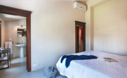 Modern bedroom interior featuring a neatly made bed with a blue throw, air conditioning unit, and en-suite bathroom access. The room is well-lit with neutral walls and wooden accents, creating a serene atmosphere.