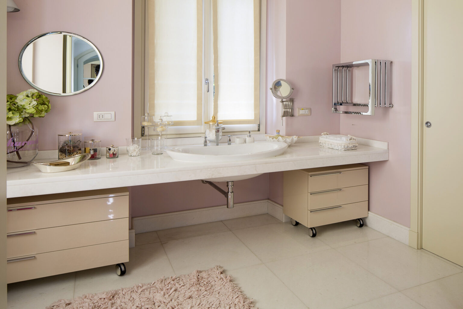 Modern bathroom interior featuring a light pink color scheme, a spacious marble countertop with a sink, stylish storage drawers on wheels, a round mirror, and elegant decor elements. Natural light floods the space through a large window, enhancing the contemporary aesthetic. | Only on SopranoVillas