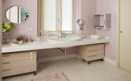 Modern bathroom interior featuring a light pink color scheme, a spacious marble countertop with a sink, stylish storage drawers on wheels, a round mirror, and elegant decor elements. Natural light floods the space through a large window, enhancing the contemporary aesthetic.