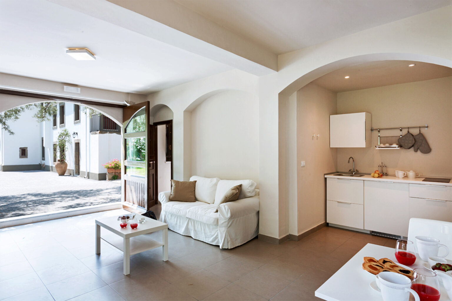 Bright and airy living space featuring a cozy white sofa, a modern kitchen area with minimalist cabinetry, and a seamless transition to an outdoor patio, perfect for relaxation and entertaining. | Only on SopranoVillas
