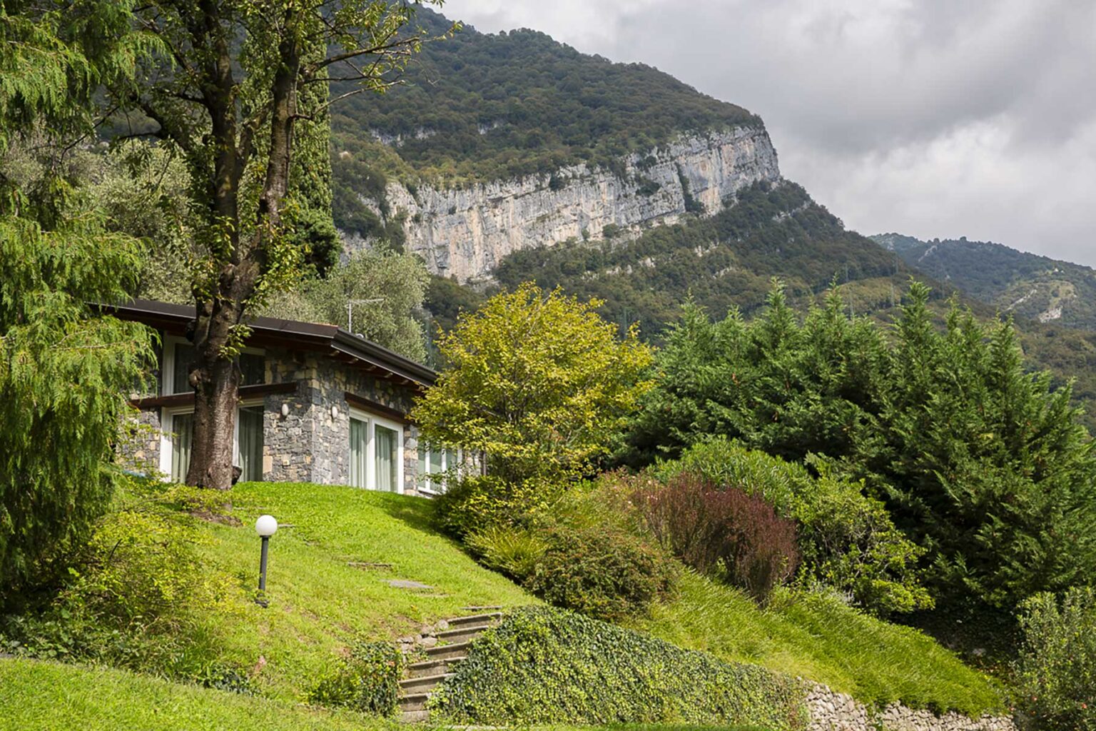 Scenic view of a stone house surrounded by lush greenery and trees, with a backdrop of dramatic mountains under a cloudy sky. Ideal for nature lovers and outdoor enthusiasts. | Only on SopranoVillas