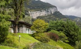 Scenic view of a stone house surrounded by lush greenery and trees, with a backdrop of dramatic mountains under a cloudy sky. Ideal for nature lovers and outdoor enthusiasts.