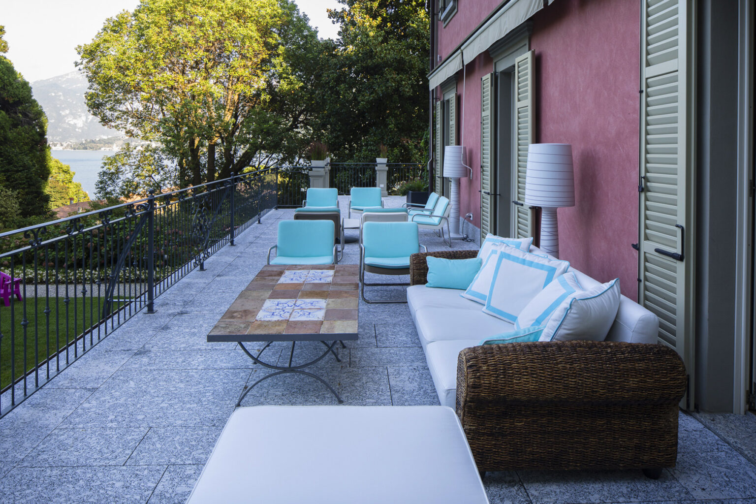 Outdoor terrace with modern seating and a wooden coffee table, surrounded by lush greenery and a scenic view. The area features white couches with turquoise accents and stylish lamps, perfect for relaxation and social gatherings. | Only on SopranoVillas