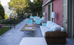 Outdoor terrace with modern seating and a wooden coffee table, surrounded by lush greenery and a scenic view. The area features white couches with turquoise accents and stylish lamps, perfect for relaxation and social gatherings.