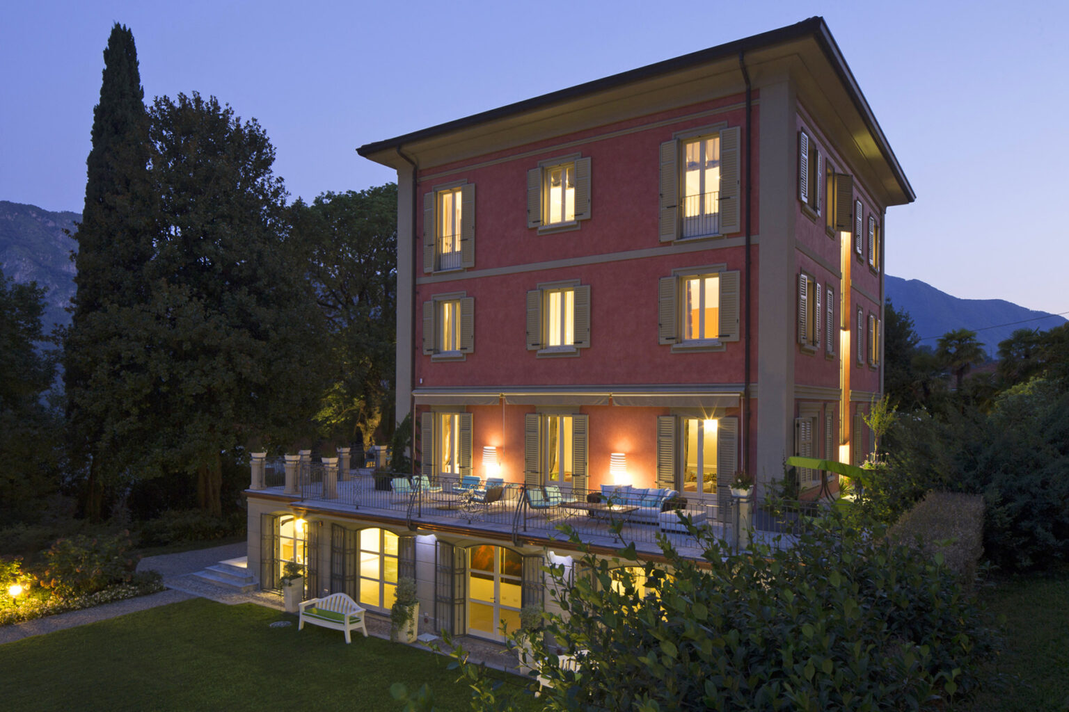 Elegant three-story red villa at dusk, featuring illuminated windows and a spacious outdoor terrace surrounded by lush greenery and mountains in the background. Ideal for luxury getaways or vacation rentals. | Only on SopranoVillas