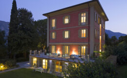 Elegant three-story red villa at dusk, featuring illuminated windows and a spacious outdoor terrace surrounded by lush greenery and mountains in the background. Ideal for luxury getaways or vacation rentals.