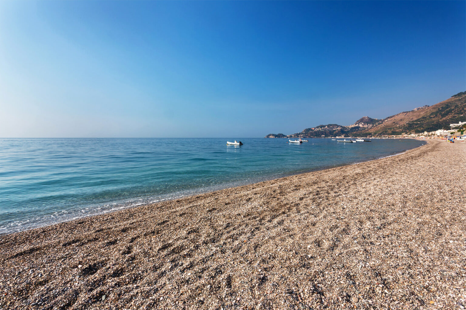 Scenic beach view with clear blue water and gentle waves, featuring a pebbled shoreline and distant boats under a bright blue sky. Ideal for vacation and relaxation. | Only on SopranoVillas