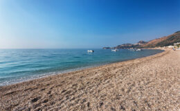 Scenic beach view with clear blue water and gentle waves, featuring a pebbled shoreline and distant boats under a bright blue sky. Ideal for vacation and relaxation.