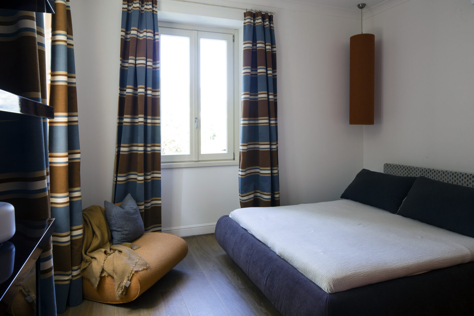 Cozy bedroom featuring a minimalist design, a comfortable bed with a light-colored mattress, and stylish curtains with a blue and brown pattern. Natural light streams in through a window, enhancing the inviting atmosphere. A plush floor cushion adds additional seating and comfort. | Only on SopranoVillas