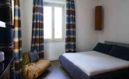Cozy bedroom featuring a minimalist design, a comfortable bed with a light-colored mattress, and stylish curtains with a blue and brown pattern. Natural light streams in through a window, enhancing the inviting atmosphere. A plush floor cushion adds additional seating and comfort.