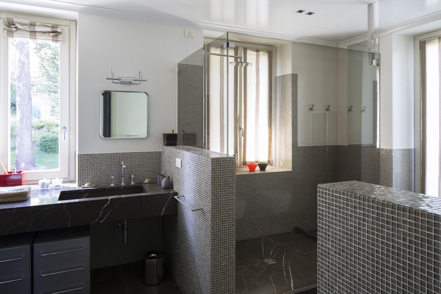 Modern bathroom interior featuring a glass shower enclosure, sleek dark cabinetry, and natural light streaming through large windows. The walls are adorned with stylish gray tiles, creating an elegant and spacious atmosphere. | Only on SopranoVillas