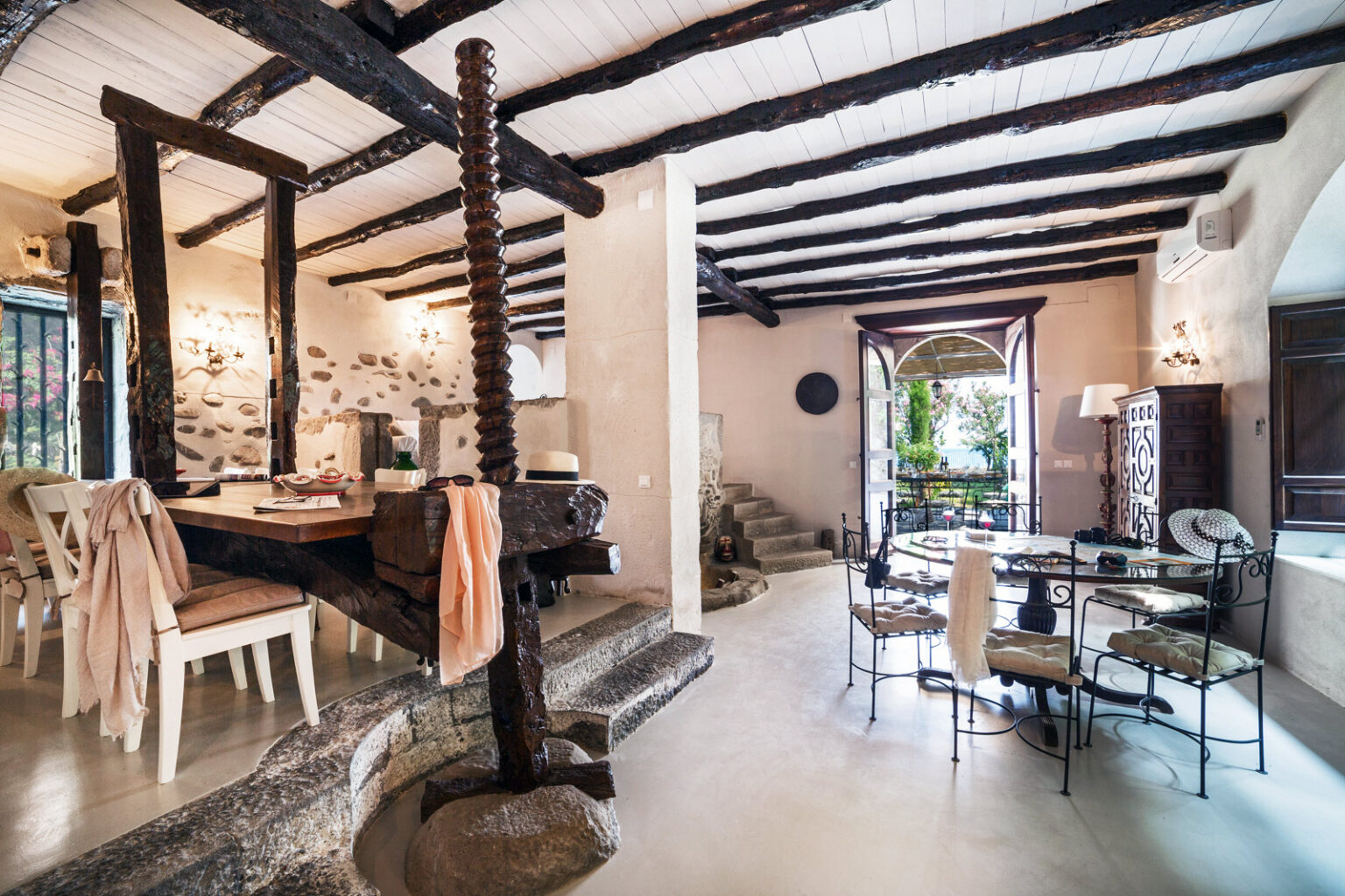 Spacious and stylish interior of a modern rustic dining and living area featuring exposed wooden beams, a stone dining table, elegant chairs, and natural light streaming through large windows. | Only on SopranoVillas