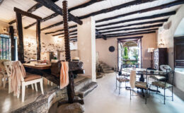 Spacious and stylish interior of a modern rustic dining and living area featuring exposed wooden beams, a stone dining table, elegant chairs, and natural light streaming through large windows.