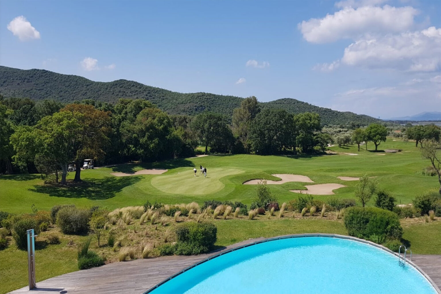 Aerial view of a lush golf course surrounded by rolling hills, featuring manicured greens and sand traps, with a swimming pool in the foreground under a clear blue sky. | Only on SopranoVillas