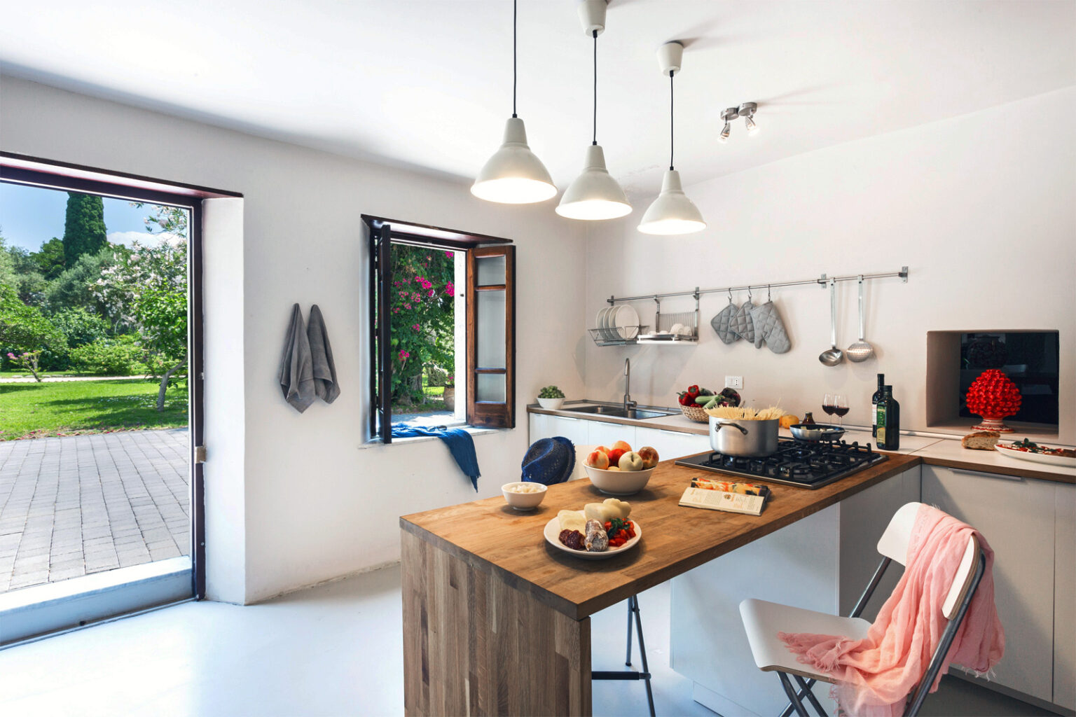 Bright and modern kitchen featuring a wooden dining table with fresh fruits, a stylish stove, and open windows leading to a lush garden. The minimalist design includes white walls, pendant lighting, and kitchen utensils neatly arranged, creating a welcoming atmosphere. | Only on SopranoVillas