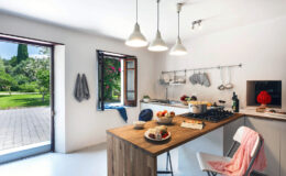 Bright and modern kitchen featuring a wooden dining table with fresh fruits, a stylish stove, and open windows leading to a lush garden. The minimalist design includes white walls, pendant lighting, and kitchen utensils neatly arranged, creating a welcoming atmosphere.