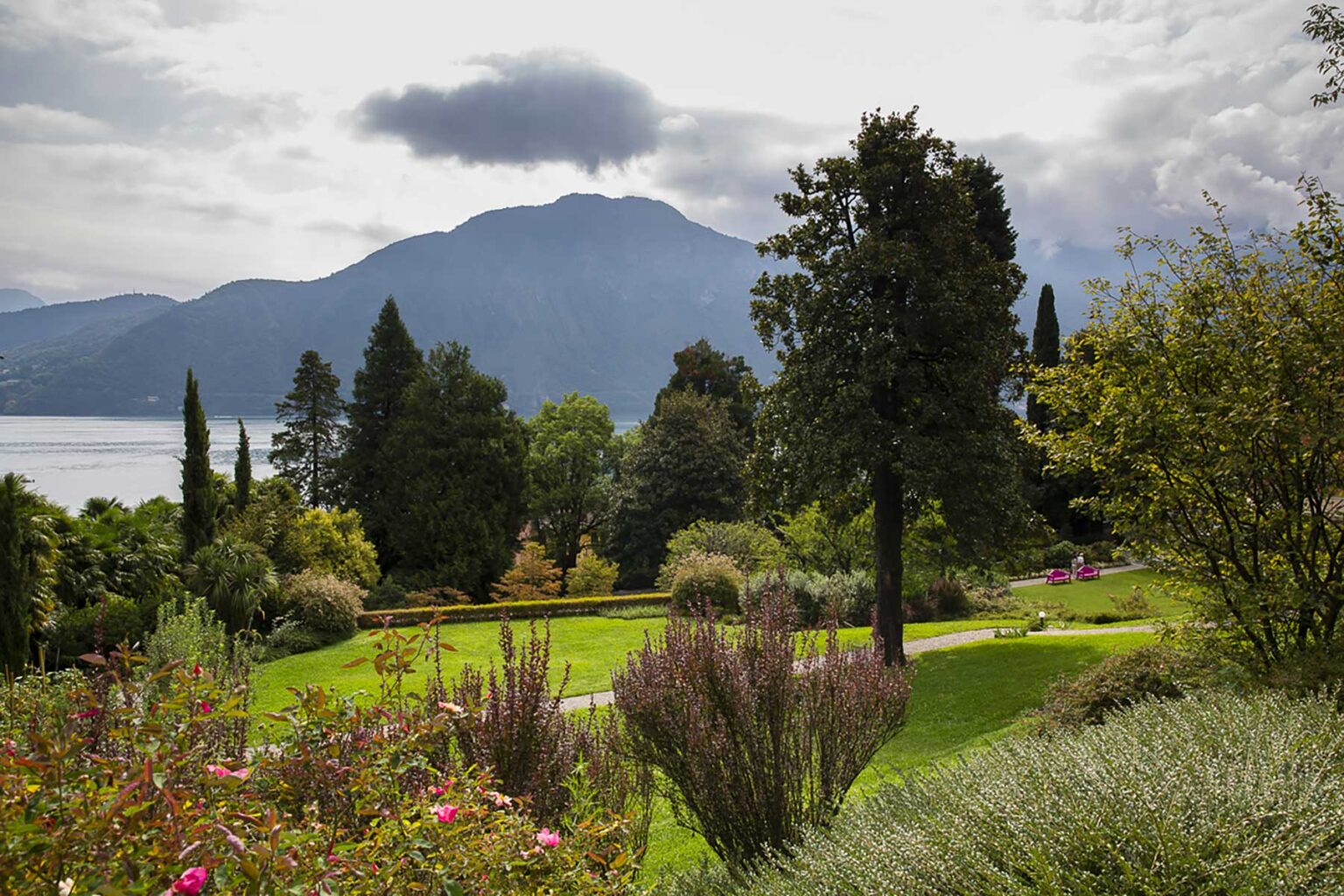 Scenic view of a lush garden with colorful flowers, trees, and a mountainous landscape in the background under a cloudy sky. | Only on SopranoVillas