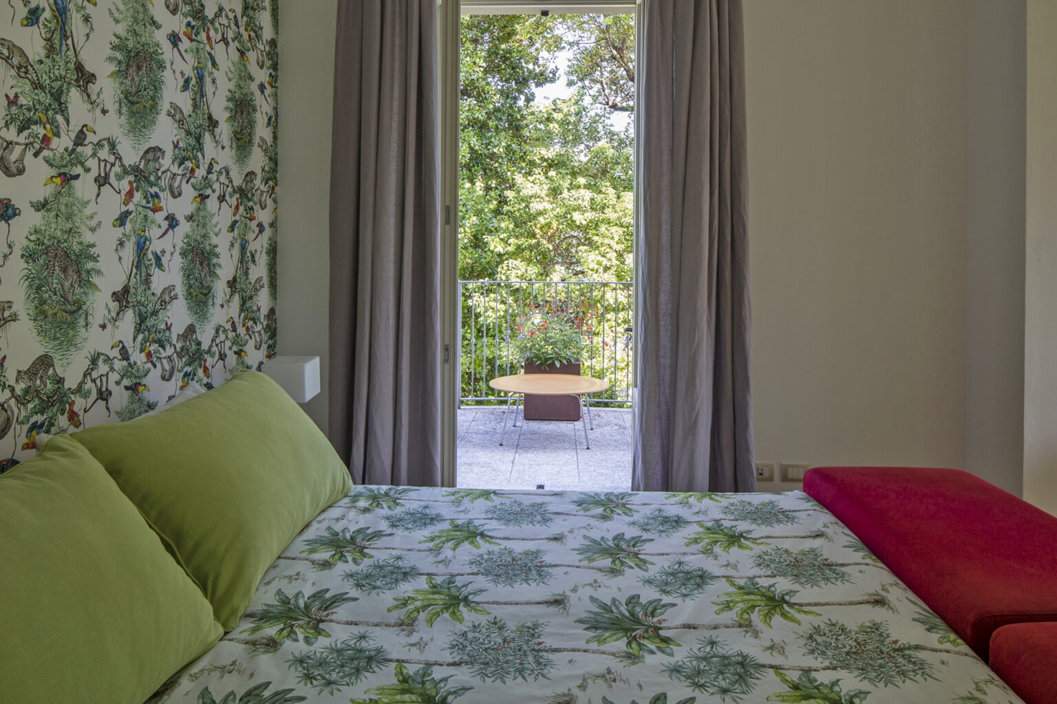 Cozy bedroom featuring a floral-patterned wall, green pillows on a bed, and a view of a balcony with outdoor seating surrounded by greenery. | Only on SopranoVillas