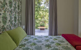 Cozy bedroom featuring a floral-patterned wall, green pillows on a bed, and a view of a balcony with outdoor seating surrounded by greenery.