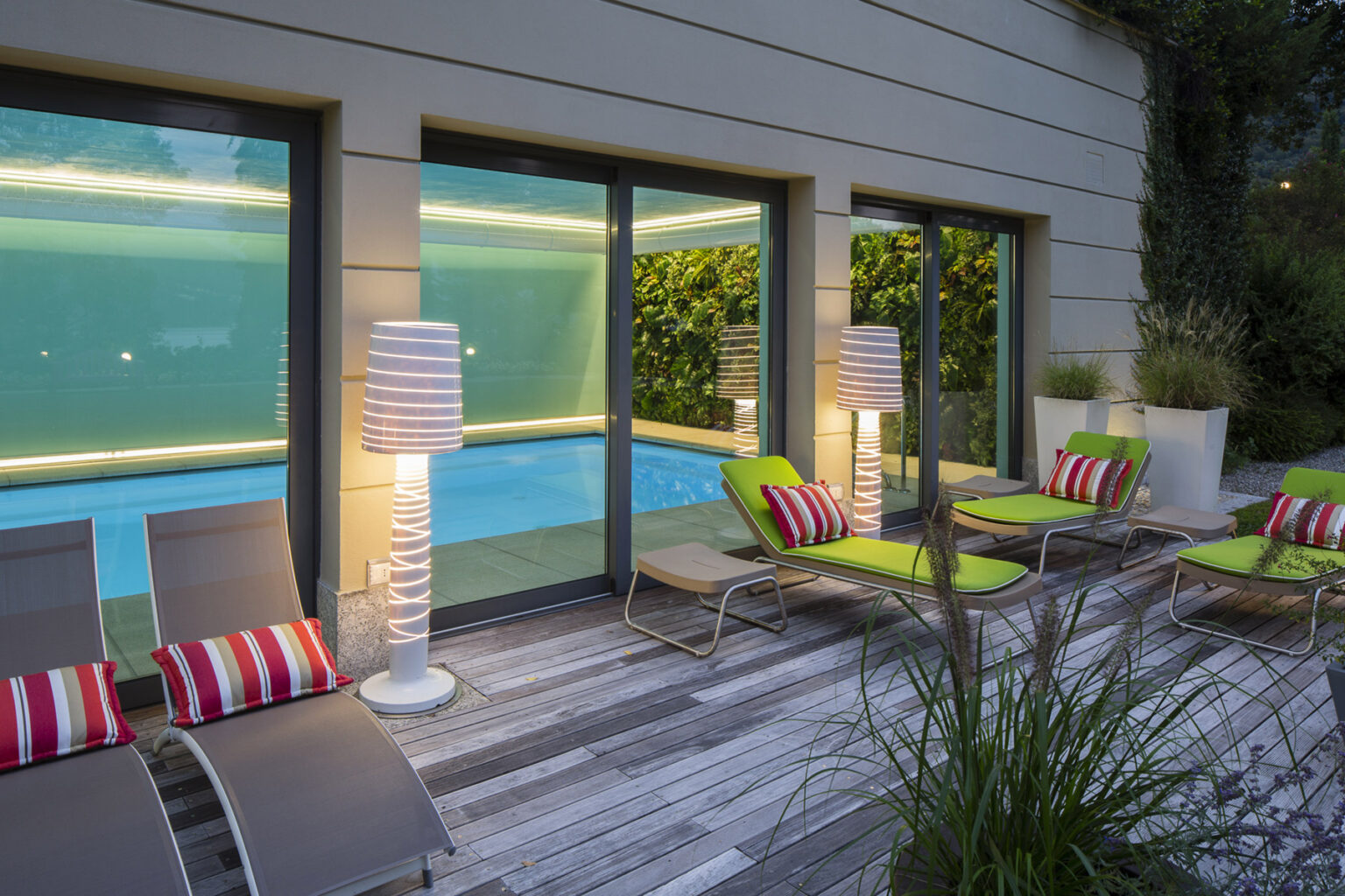 Modern outdoor lounge area featuring a swimming pool, stylish green and striped lounge chairs, and illuminated lamps, surrounded by lush greenery. | Only on SopranoVillas