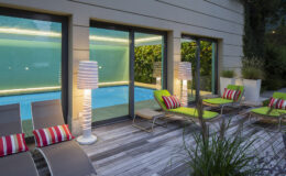 Modern outdoor lounge area featuring a swimming pool, stylish green and striped lounge chairs, and illuminated lamps, surrounded by lush greenery.