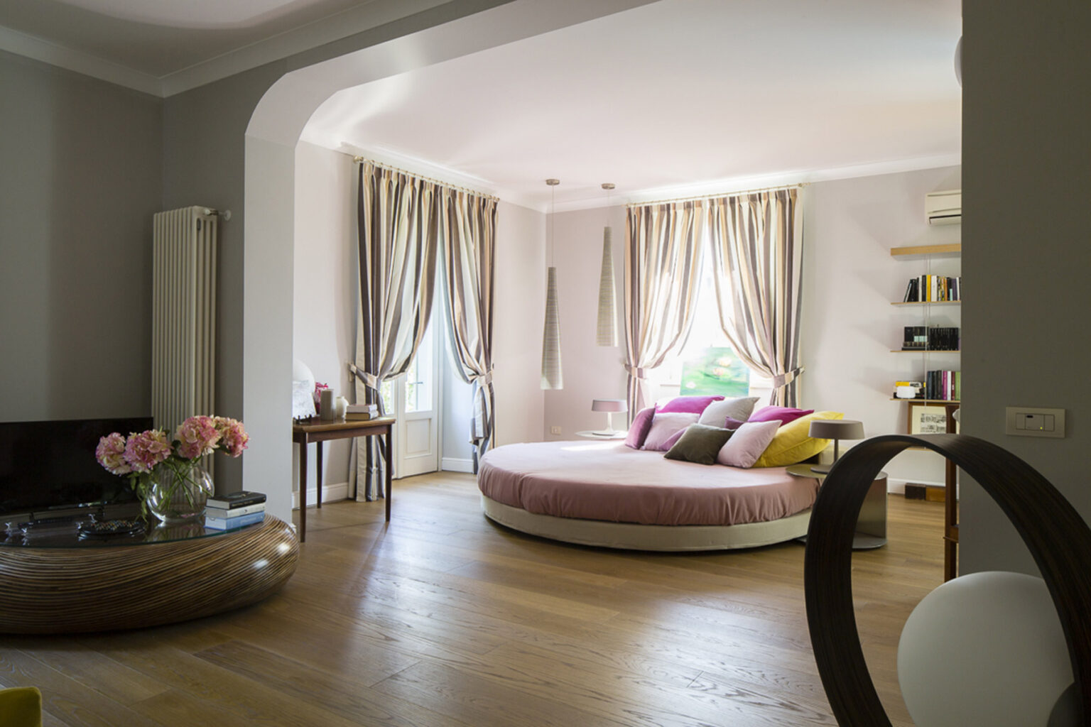 Modern living room featuring a circular bed with colorful pillows, elegant curtains, and wooden flooring. A stylish coffee table and decorative shelves add to the contemporary design. | Only on SopranoVillas