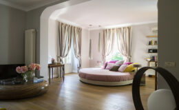 Modern living room featuring a circular bed with colorful pillows, elegant curtains, and wooden flooring. A stylish coffee table and decorative shelves add to the contemporary design.