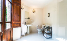 Bright and airy bathroom featuring a wooden window, a modern sink, a toilet, and a stylish storage cart. Natural light illuminates the space, highlighting the minimalist design and elegant decor elements.