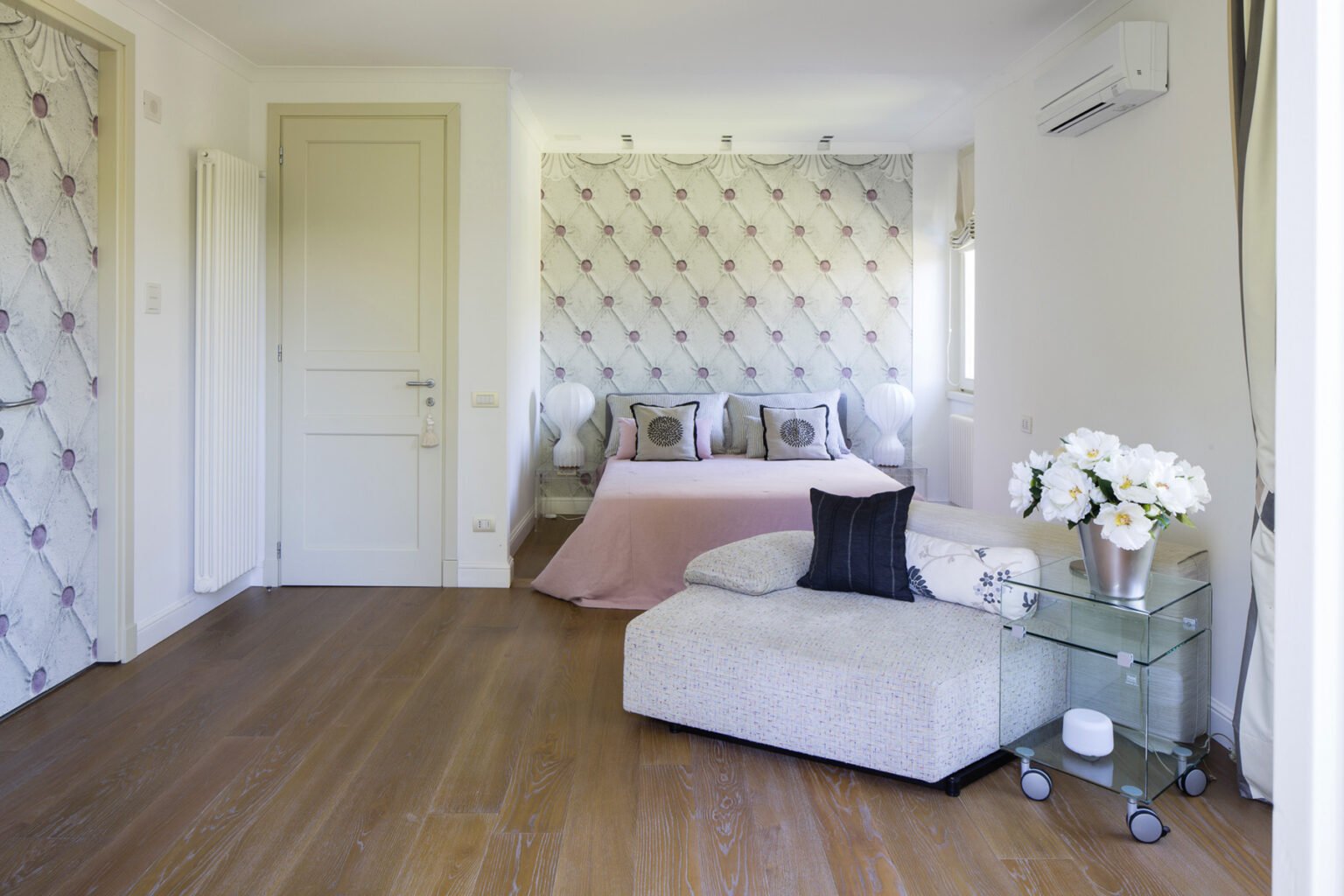 Modern bedroom interior featuring a stylish bed with decorative pillows, a cozy seating area with an ottoman, elegant wallpaper, and a floral arrangement on a glass side table, all complemented by warm wooden flooring. | Only on SopranoVillas