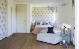 Modern bedroom interior featuring a stylish bed with decorative pillows, a cozy seating area with an ottoman, elegant wallpaper, and a floral arrangement on a glass side table, all complemented by warm wooden flooring.