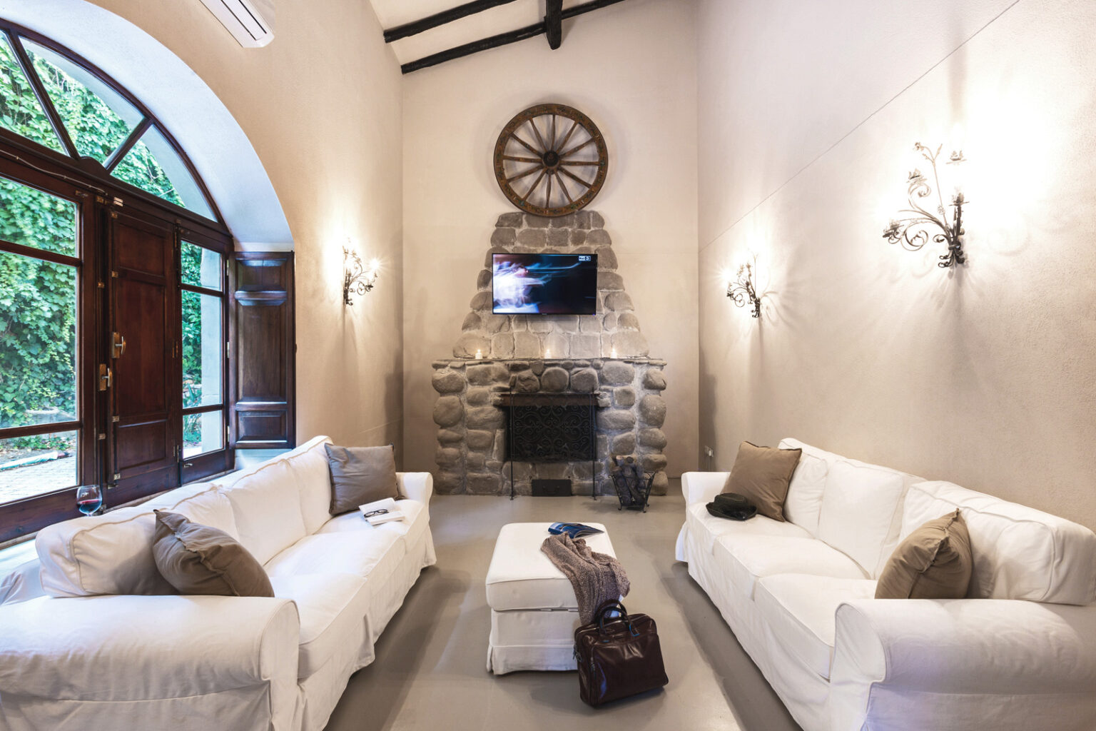 Cozy living room featuring white sofas, a stone fireplace, and a mounted TV, with large arched windows allowing natural light and a view of greenery. | Only on SopranoVillas