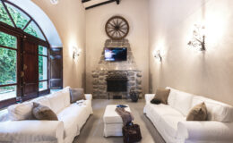 Cozy living room featuring white sofas, a stone fireplace, and a mounted TV, with large arched windows allowing natural light and a view of greenery.