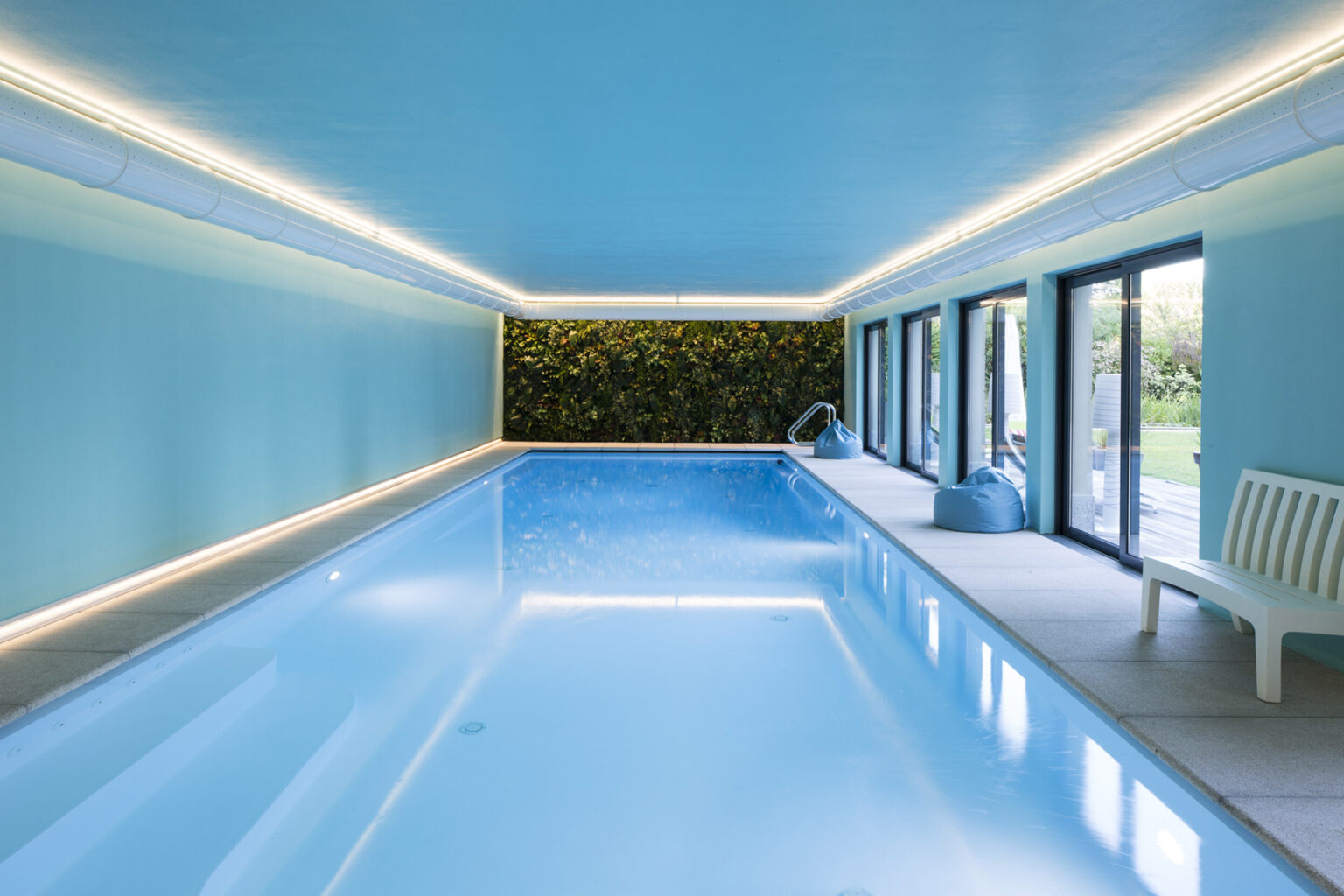 Indoor swimming pool with a light blue ceiling and walls, featuring modern LED lighting and a green plant wall. Large windows provide natural light and a view of the outdoor area, enhancing the tranquil atmosphere. | Only on SopranoVillas
