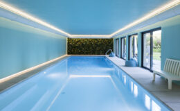 Indoor swimming pool with a light blue ceiling and walls, featuring modern LED lighting and a green plant wall. Large windows provide natural light and a view of the outdoor area, enhancing the tranquil atmosphere.