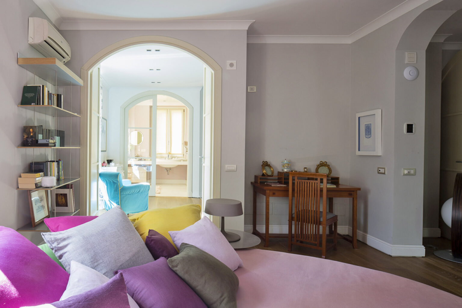 Cozy interior view of a modern living space featuring a pink sofa with colorful pillows, a wooden desk, and a bright archway leading to a well-lit room with colorful chairs and shelves. The walls are painted in soft gray tones, enhancing the inviting atmosphere. | Only on SopranoVillas