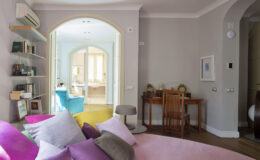 Cozy interior view of a modern living space featuring a pink sofa with colorful pillows, a wooden desk, and a bright archway leading to a well-lit room with colorful chairs and shelves. The walls are painted in soft gray tones, enhancing the inviting atmosphere.
