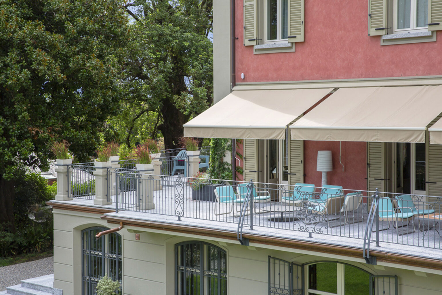 Modern balcony with turquoise chairs and a white table, overlooking a garden, featuring beige awnings on a pink building. Ideal for outdoor relaxation and entertaining. | Only on SopranoVillas
