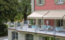 Modern balcony with turquoise chairs and a white table, overlooking a garden, featuring beige awnings on a pink building. Ideal for outdoor relaxation and entertaining.