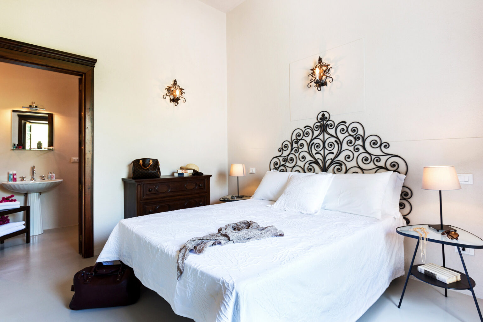 Cozy bedroom featuring a large white bed with an ornate black headboard, soft lighting from bedside lamps, and a stylish nightstand. The room includes a doorway leading to a modern bathroom, creating a welcoming and serene atmosphere. Ideal for relaxation and comfort. | Only on SopranoVillas