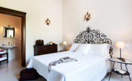 Cozy bedroom featuring a large white bed with an ornate black headboard, soft lighting from bedside lamps, and a stylish nightstand. The room includes a doorway leading to a modern bathroom, creating a welcoming and serene atmosphere. Ideal for relaxation and comfort.