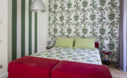 Cozy bedroom featuring a floral wallpaper accent wall, green bedding, and a red ottoman, creating a vibrant and inviting atmosphere.