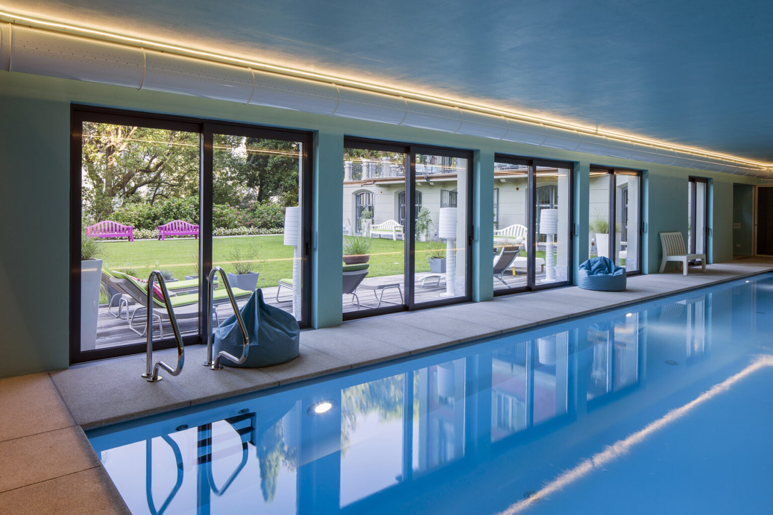 Indoor swimming pool with large windows overlooking a landscaped garden, featuring modern lounge chairs and a stylish ambiance. | Only on SopranoVillas