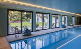 Indoor swimming pool with large windows overlooking a landscaped garden, featuring modern lounge chairs and a stylish ambiance.