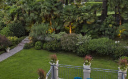 Lush garden view featuring manicured lawns, tropical plants, and a stone pathway, with decorative planters and outdoor seating visible in the foreground.