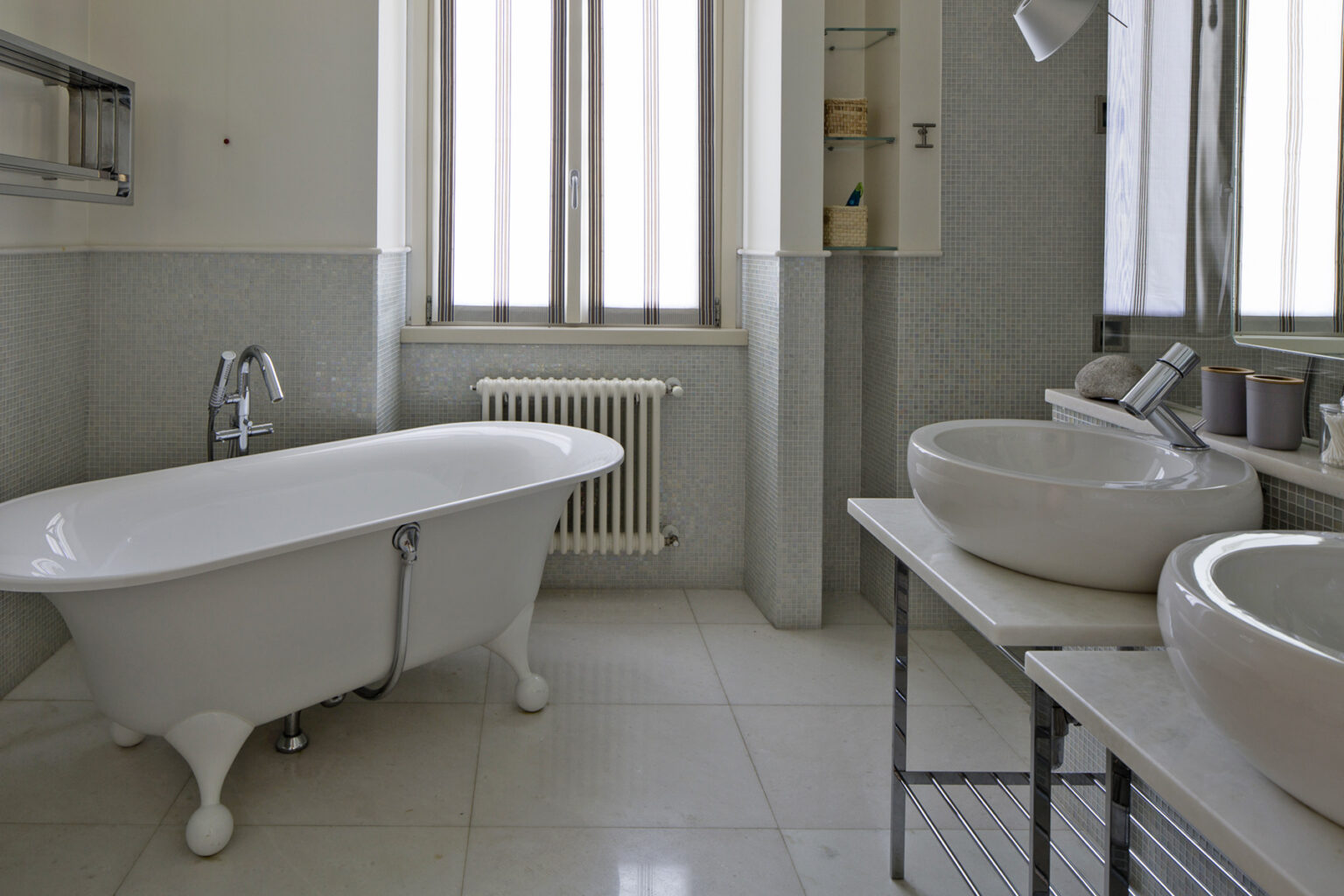 Modern bathroom featuring a freestanding white bathtub, two stylish sinks on a metal frame, and large windows providing natural light. The walls are finished in light gray tiles, creating a serene atmosphere. | Only on SopranoVillas