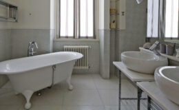 Modern bathroom featuring a freestanding white bathtub, two stylish sinks on a metal frame, and large windows providing natural light. The walls are finished in light gray tiles, creating a serene atmosphere.